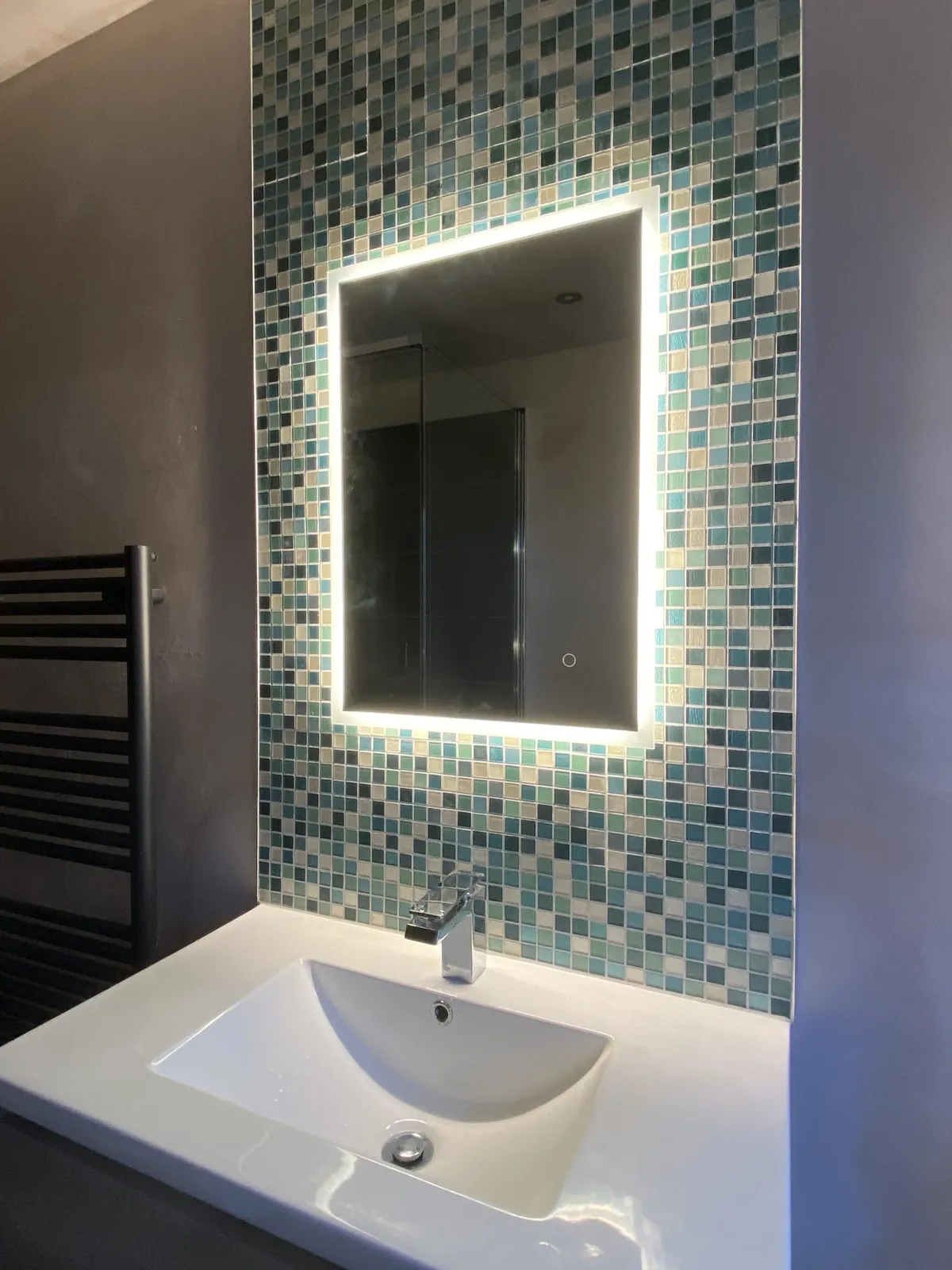 Glass mosaic tile wall behind a lit bathroom mirror and white sink